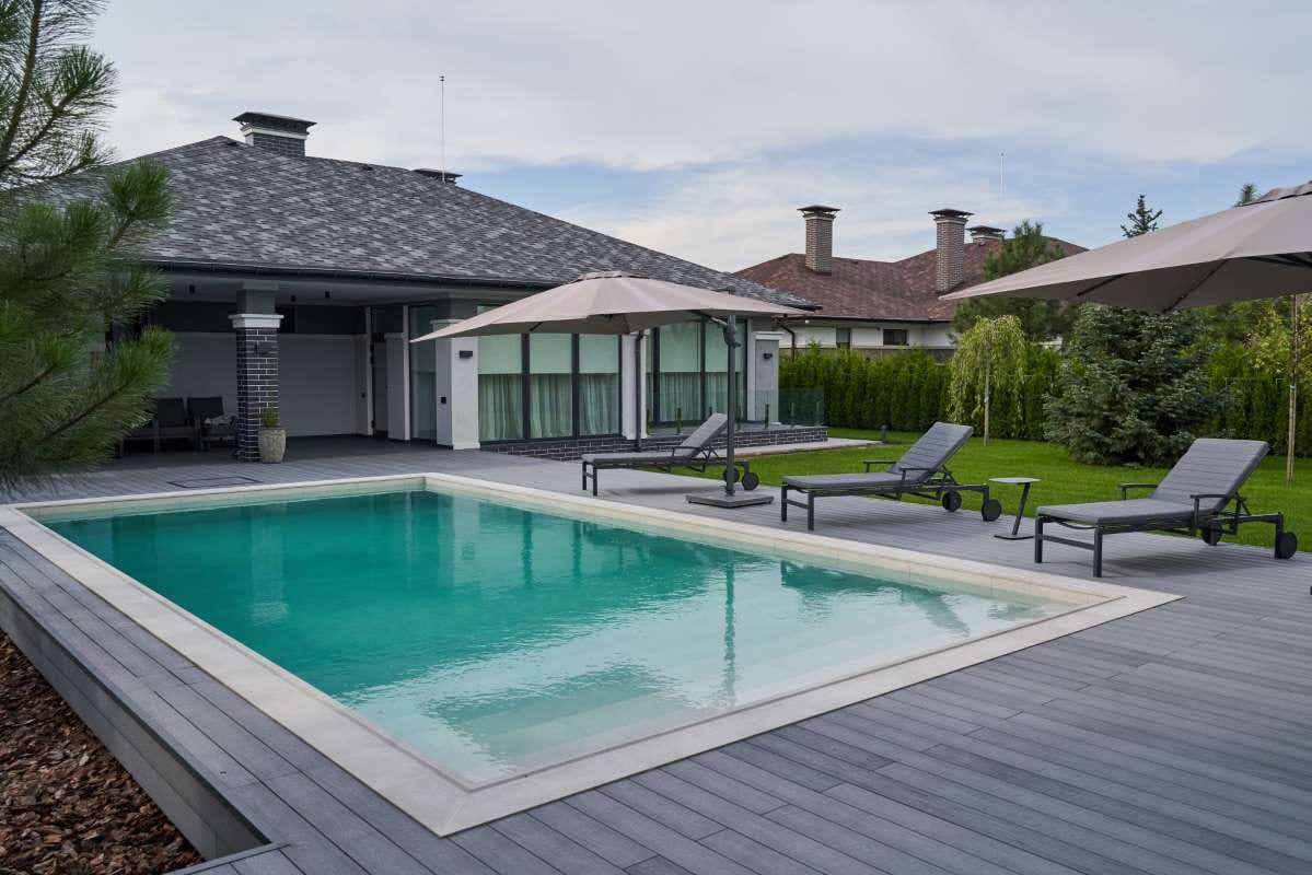 Modern villa pool deck with wooden lounge chairs and light blue water